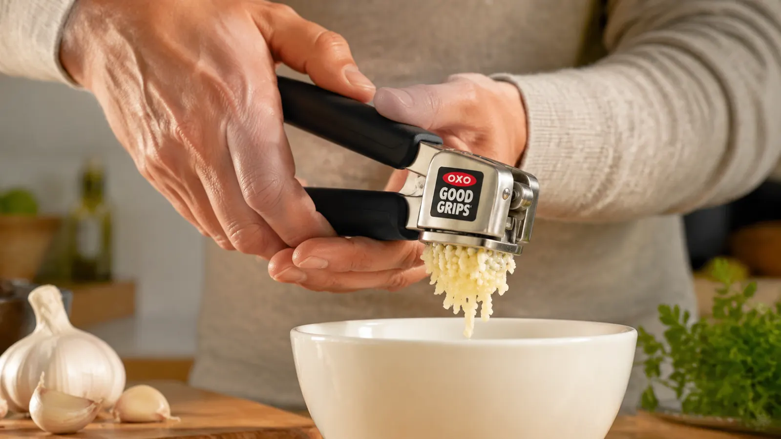 Hand squeezing the OXO garlic press handles together over a small ceramic prep bowl, minced garlic coming through the hopper