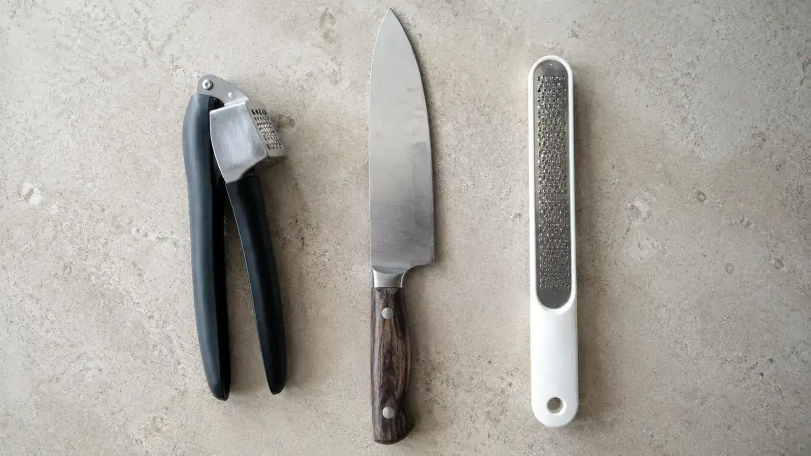 Lineup of three garlic preparation tools on a kitchen counter: OXO garlic press, a chef's knife, and a ceramic microplane grater