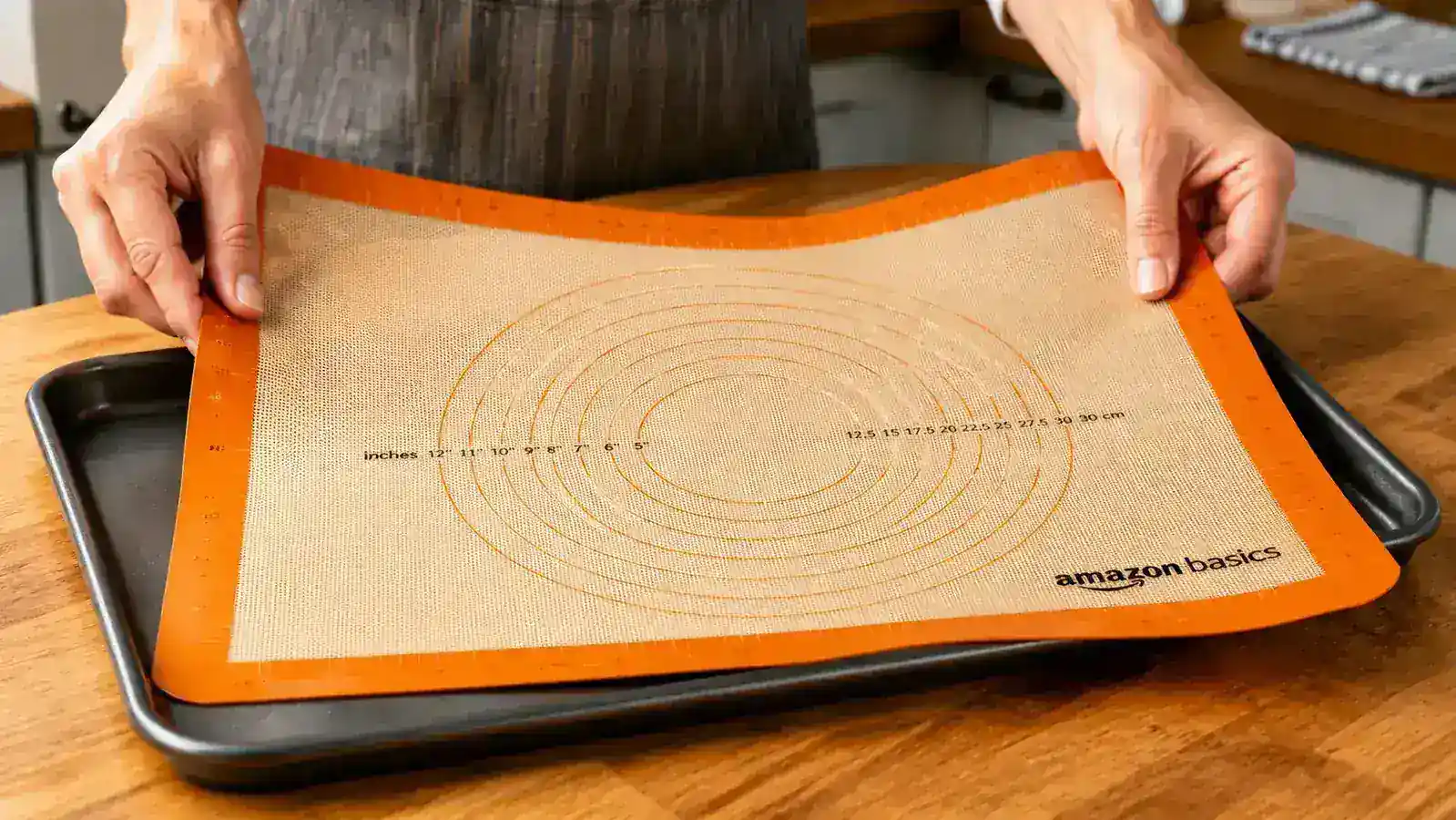 Hands unrolling a silicone baking mat onto a dark baking sheet on a wooden countertop